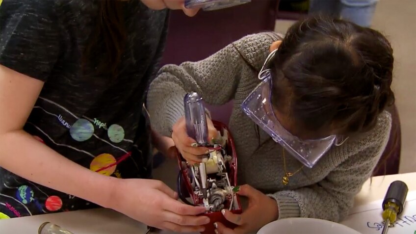 A young girl works on a piece of machinery as another child helps her hold it.