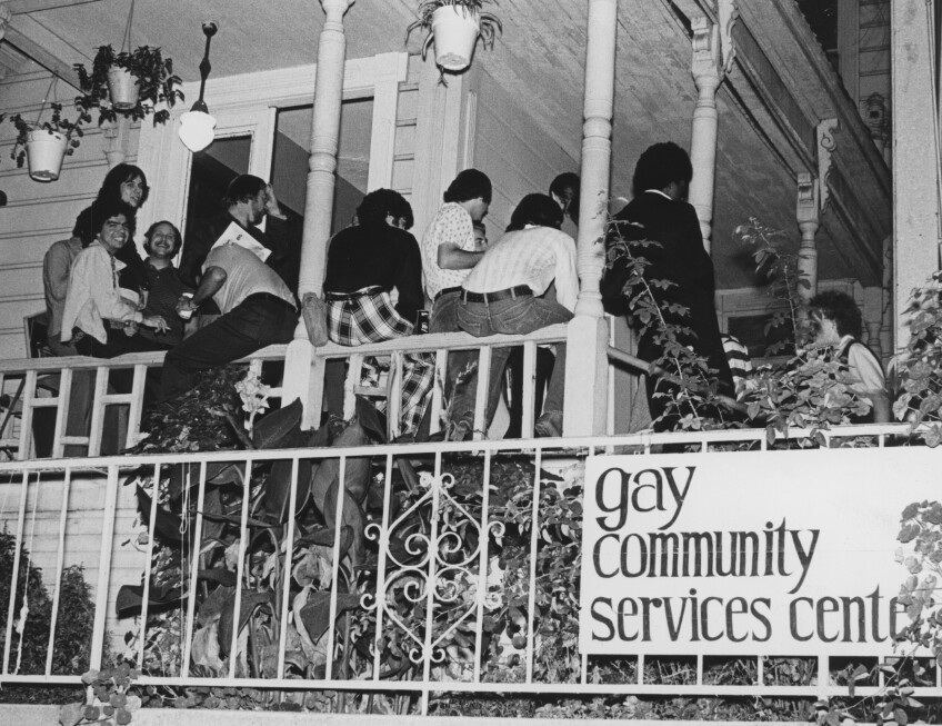 A black and white photo of young people gathered on a house porch. Many are sitting on the railing. Below, foliage and plants are fenced in with an iron-wrought fence. On the fence a sign that reads, "Gay Community Services Center" is hung.   