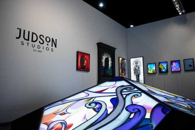 Glass artworks displayed on white gallery walls. At the foreground is a glass artwork shaped as a pointed dome with colored glass shapes. On the gallery wall behind reads,