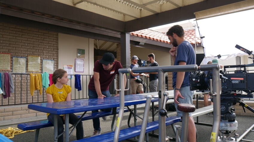 A young girl in a yellow t-shirt is seated a blue table in an outdoor elementary school setting. Next to her, two people are standing in front of and speaks to her.  