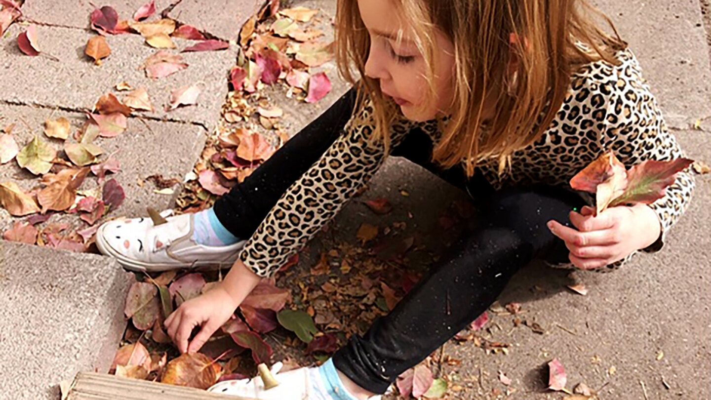 A small blonde girl sits on the floor gathering leaves