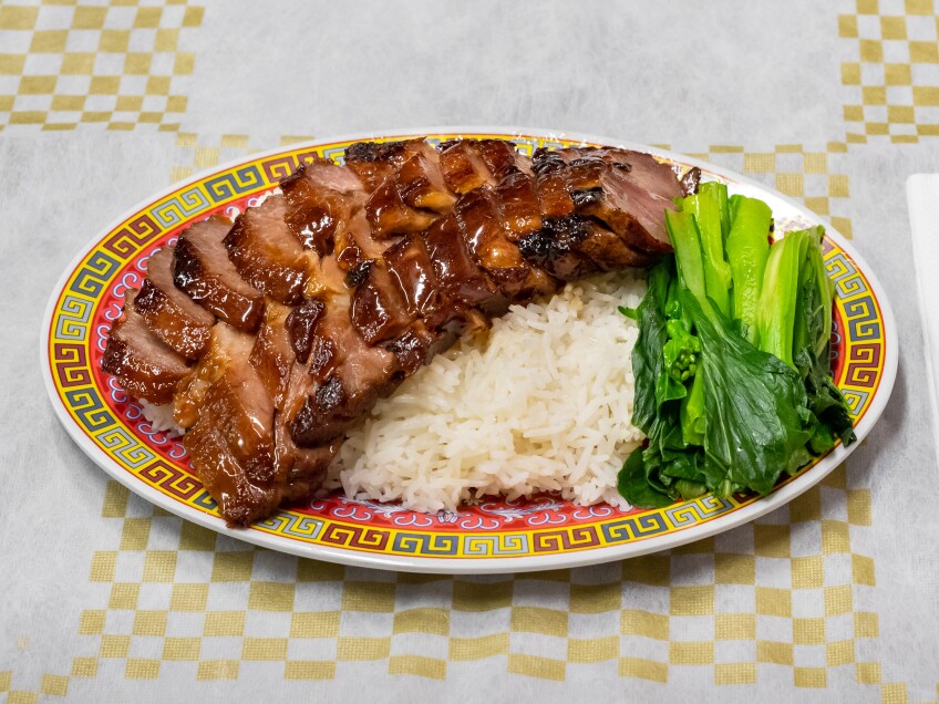 A plate of red roasted char siu served over a bed of fluffy white rice and a side of leafy greens. The plate is set atop a yellow and white checkered tablecloth.