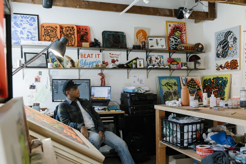 DJ Javier sits in front of a desk, facing his art studio. The studio walls are covered with artwork hung on the wall and displayed on floating shelves. In the center of the studio is a wooden work table topped with art supplies and materials.