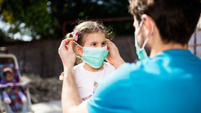A grown-up puts a face mask on a small child while outside.