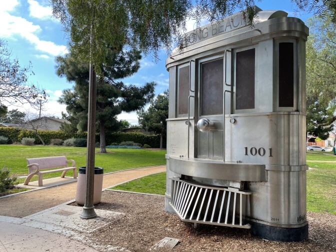 A stainless steel replica of a Red Car trolley stands in a lush, green park. On the trolley replica are the numbers, "1001." At the top of the replica is a sign that reads, "Long Beach."