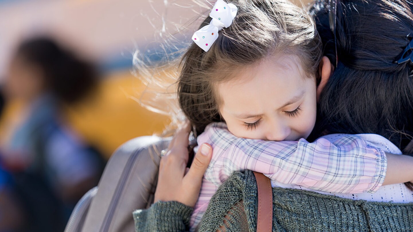 A small child hugs a caregiver goodbye in front of a school bus.