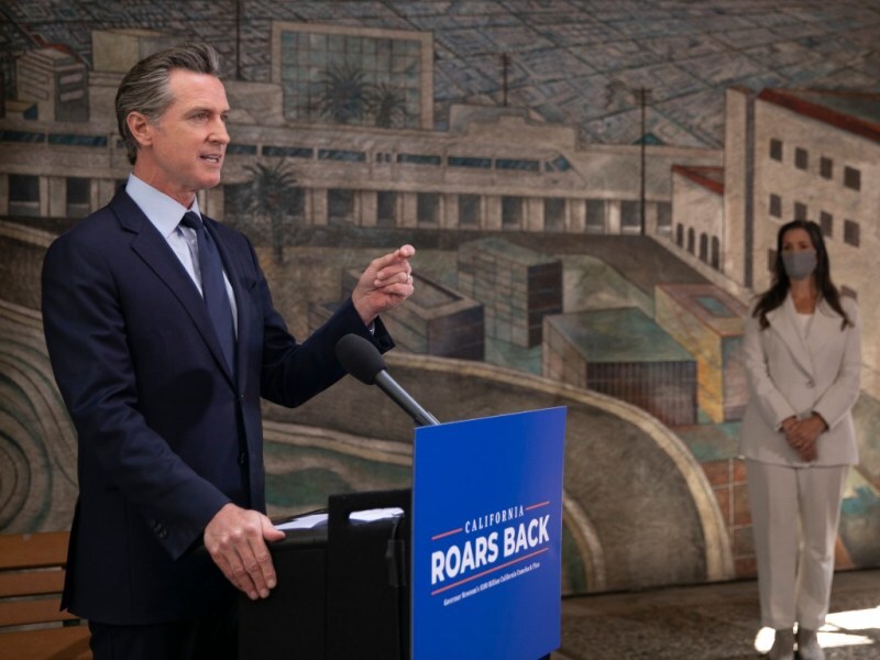 Photo of Governor Gavin Newsom standing at a podium with a poster that says "California Roars Back". There is a mural in the background.