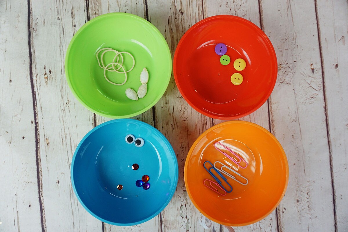 Cuatro tazones de colores llenos de artículos ordenados por tipo, incluyendo ligas, semillas, clips de papel, botones y lentejuelas.