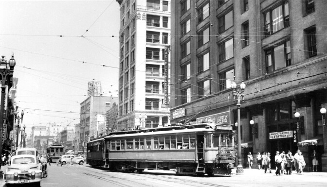 An old black and white photo of aa Pacific Electric Railway station. A red car approaches the terminal. Exposed tracks and a tangle of wires above the street are reminiscent of the red car heyday.