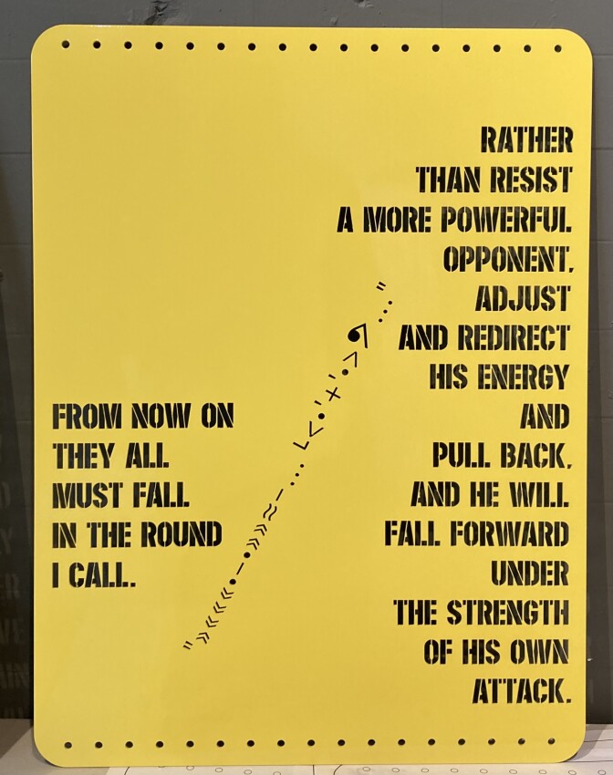 A yellow board that has stenciled words which read "From now on, they all, must fall, in the round, I call" and "Rather, than resist, a more powerful, opponent, and redirect, his energy, and, pull back, and he will, fall forward, under, the strength, of his own, attack."