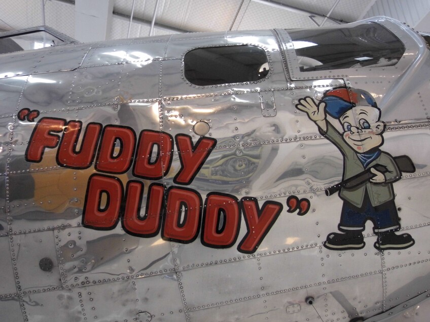 The side of a metal aircraft with the words, "Fuddy Duddy" painted in red next to a cartoon character of a man holding a rifle. 