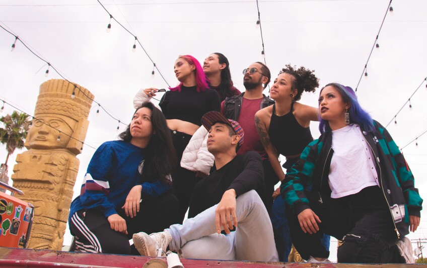 Members of Temper Tantrum huddle, with some members sitting, some kneeling, some crouched down and some standing up. They are all looking off to something out of frame to the left. They are dressed in casual streetwear in an outdoor setting. Electric string lights hang above them.
