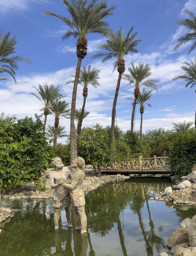 A statue of two people dancing stands in the middle of a reflective pond. The water is so still that it reflects the shrubbery, date palms, and blue sky above. Beyond the statues a small, wooden footbridge crosses over a narrow part of the pond. Tall date trees tower of the scene.