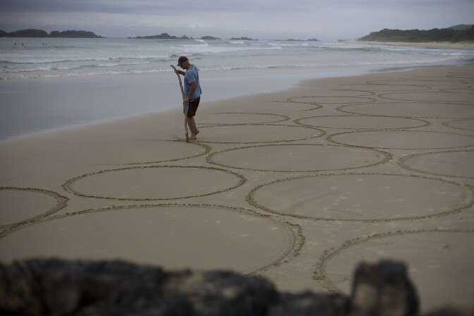 A man in a blue t-shirt and a black cap draws large perfect circles onto the sand, using a tree branch. He's surrounded by several other circles of the same size. Nearby is a shoreline, lapping at the coast and erasing parts of the design.