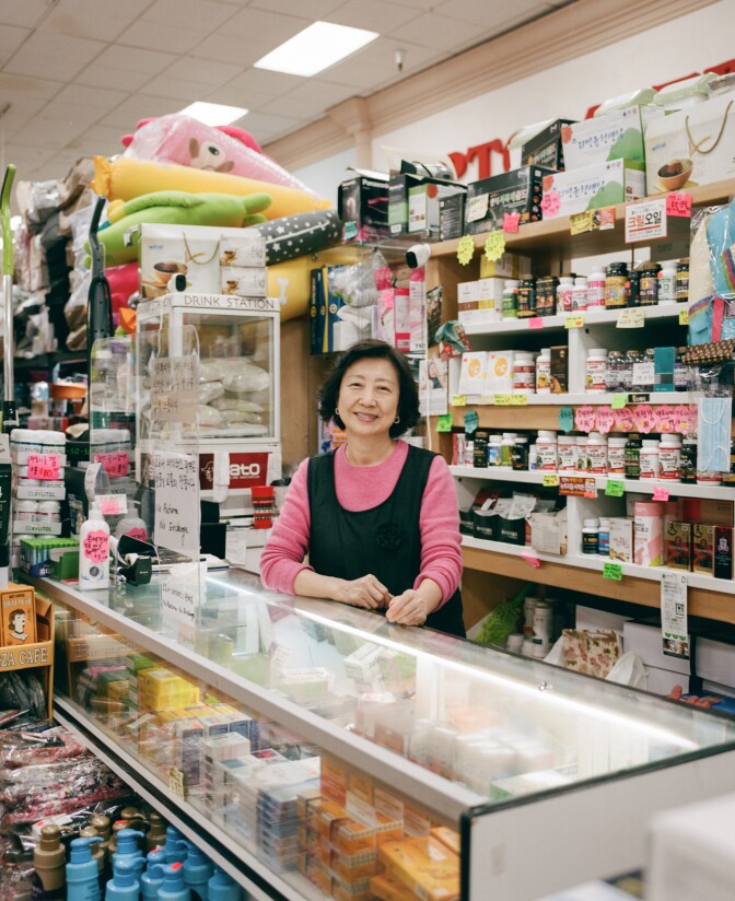 A shopkeeper in Koreatown stands by the checkout counter smiling with rows of goods behind her.