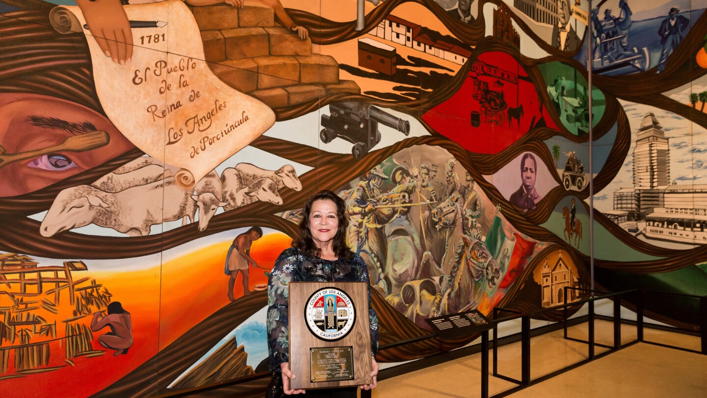 Artist Barbara Carrasco standing in front of colorful floor-to-wall length mural "L.A. History: A Mexican Perspective" holding a commendation plaque from Los Angeles County presented by County Supervisor Hilda Solis at the opening of the temporary exhibition Sin Censura A.