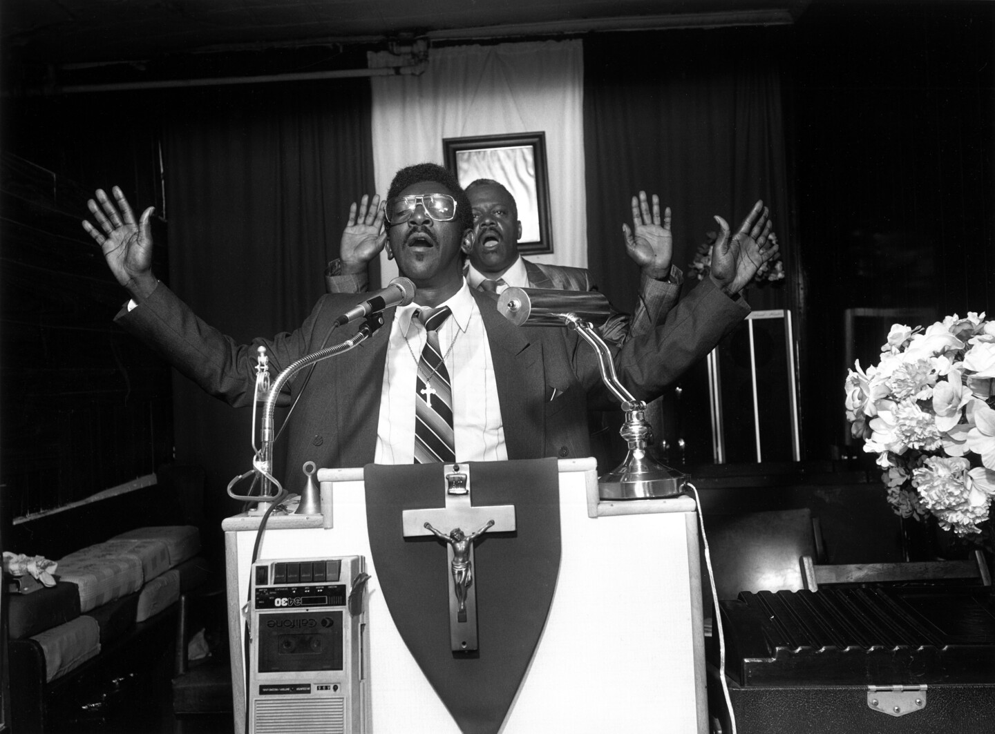 Two men, preaching, arms outstretched with Jesus Christ on a cross plaque hanging on the podium.