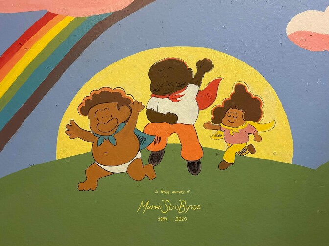 A mural depicts a Black family running in a vast green landscape with a rainbow and sun shining brightly behind them. The three are wearing capes and have joyful expressions on their faces.