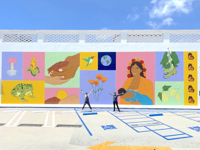 Two women stand in front of a large and colorful mural. The mural consists of images segregated in their own square, each with their own color block background. One of the squares is yellow with a frog. Another square is pink with the image of a dark-skinned woman with long hair and holding a baby. Other imagery on the tiles include California poppies, corn, a hummingbird, the earth, butterflies and two hands holding soil.