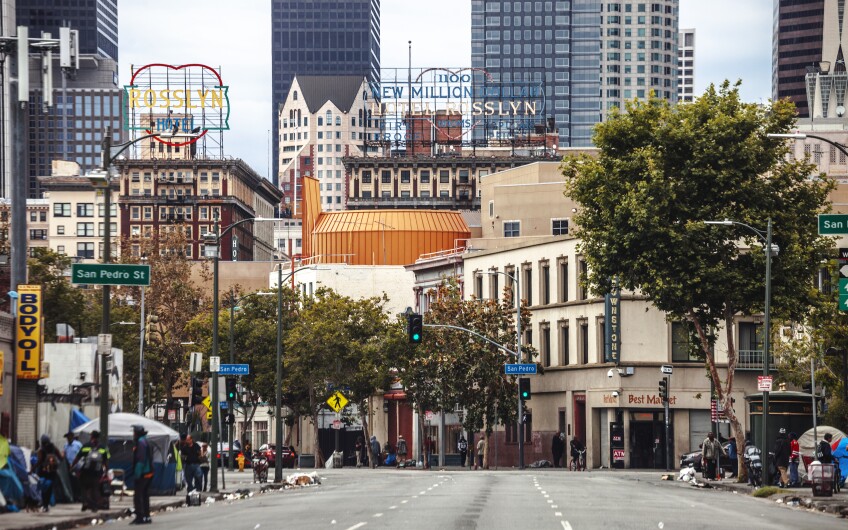 A view looking down Skid Row at San Pedro Street with the DTLA skyline, including the Rosslyn Hotel, in the distance and homeless encampments in the foreground.