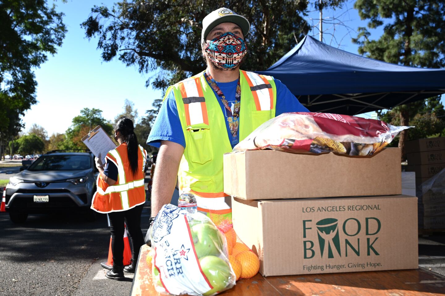 A man in a bright yellow reflective safety vest stands behind two boxes stacked on a table. There are also bags of fruit. The man is wearing a mask and looking off to the distance. Behind him is a person wearing a bright orange safety vest and holding a clipboard. Their back is facing the camera and it appears as if they are guiding a line of cars.