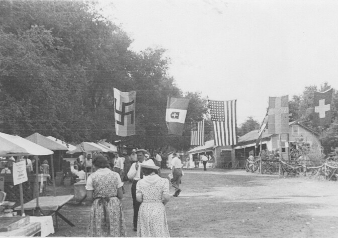 A black and white archival photo of a park with large flags draped across an empty field. The left most flag has a Swastika on it. Booths are set up along a tree line and attendees are spaced out in the park.