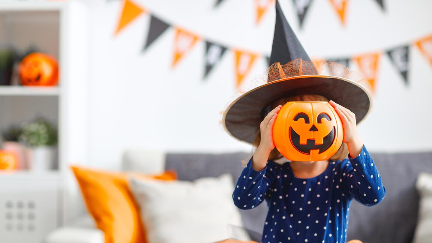 A little girl in a witch hat holds a plastic jack o' lantern over her face.