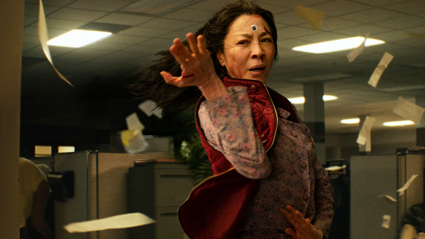 Michelle Yeoh is in a warrior pose, both hands in a pushing in one direction, palms open. Her hair is flying in a gust of wind that is also blowing various papers that swirl around her. She is in a dimly lit office floor with rows of cubicles. Yeoh's character has blood smeared across her forehead and dripping down one nostril. A googley eye is stuck on her forehead, between her brows.