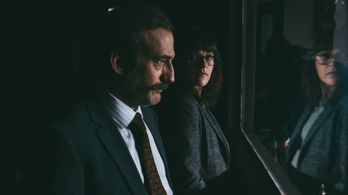 Two people stand at the other side of a one-way mirror in an interrogation room.