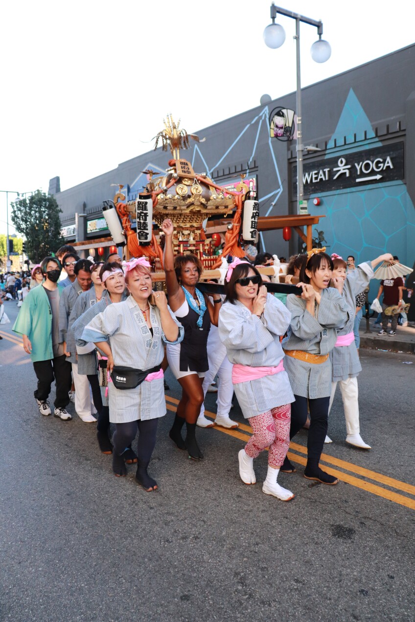 Parade participants carry a portable miniature shrine on their shoulders as they walk down the street. Women lead the group at the front, wearing gray kimonos. Onlookers watch from the curb. 
