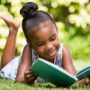 A small child smiles as she reads a book in a grassy place.