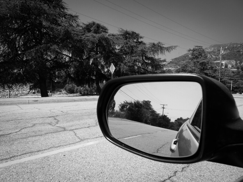 A black and white photo of a car's side view mirror, viewing the curve of a road's downward slope. Beyond the side view mirror is a road, trees and a mountain.