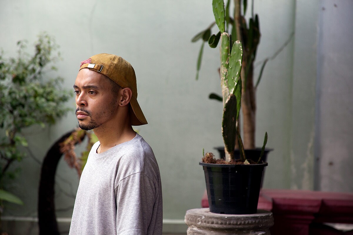 Dancer Jay Carlon poses in a white shirt and mustard cap near some potted plants at Bangkok 1899. | Matthieu Lunard