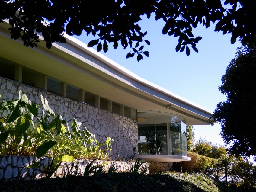 A flat and low-ceiling modernist home with a cobble-stoned wall and floor to ceiling glass windows. Surrounding the home are trees with large and long branches. At the side of the home are beds of tropical plants.  
