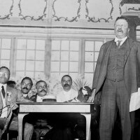 Theodore Roosevelt (foreground) stands on stage before an audience with Booker T. Washington (right) sitting behind him. | Still from "City Rising: The Informal Economy"