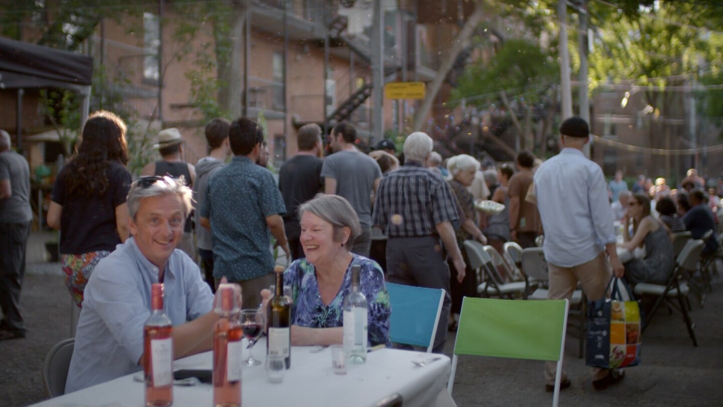 Mikael Coville-Andersen sits with a woman at a table and smiles at the camera, while a large gathering of people stand and sit at tables behind them. | From "Life-Sized City: Montreal"