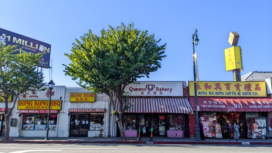 Three storefronts along a main road. The store on the left is a restaurant, Hong Kong BBQ. The store in the middle is Queen's Bakery. And the store on the right is Wing Wa Hing Gifts & Arts Co.