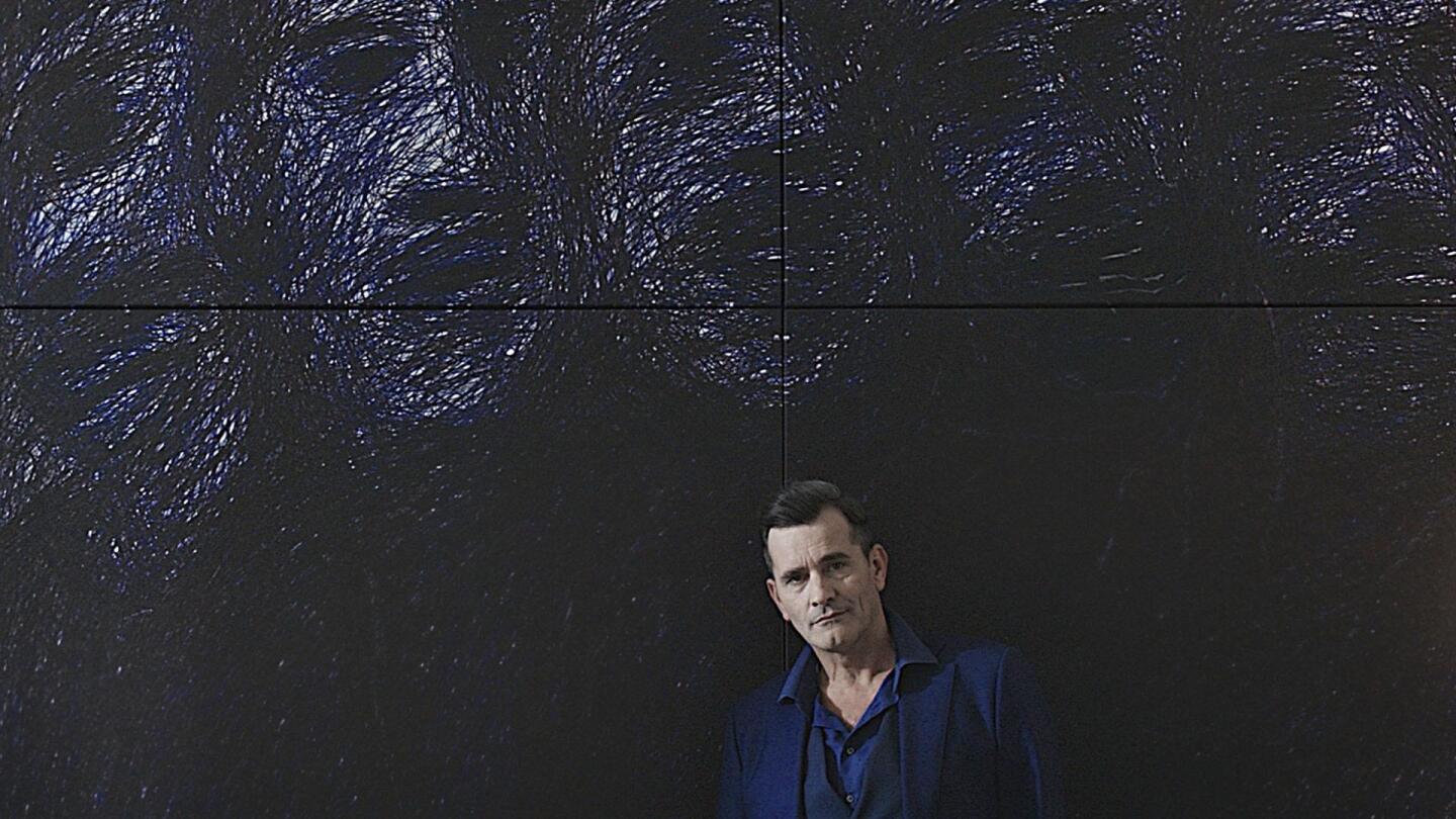 Professor T (Koen De Bouw) standing against a dark wall with impressionistic images of faces on it. | "Professor T: Season 2 - Diamond"
