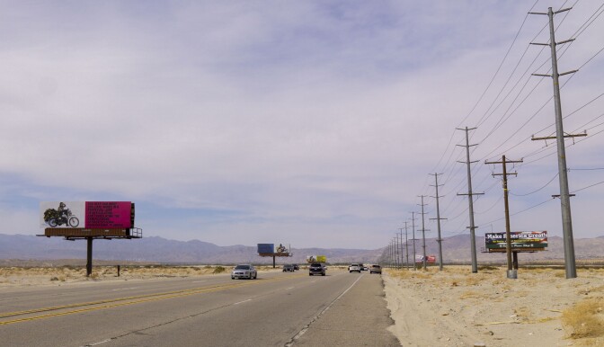 Simmons' "Because You Know Ultimately We Will Band a Militia" billboard art installation stands opposite of a billboard advertisement for Rocky's Pawn Shop. Behind the billboards is a clear, landscape view of the desert. Cars drive on the road that separates the installation and advertisement.