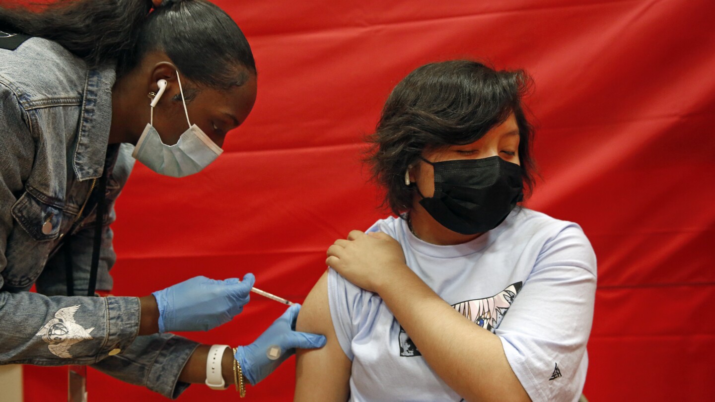 A young man in long black hair gets a vaccine from a young Black woman wearing a ponytail.