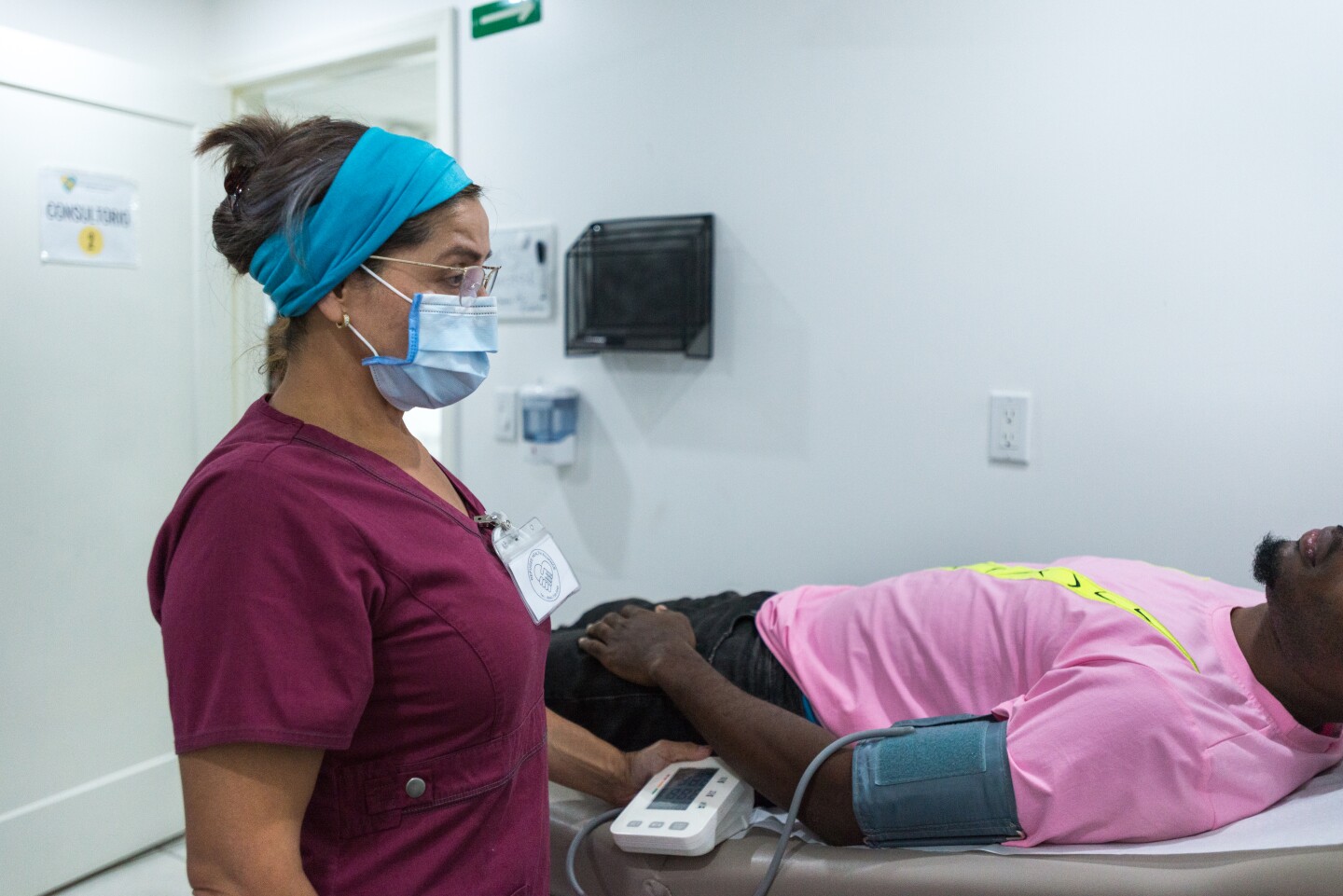 A nurse stands over a man laying on a hospital bench. He's wearing a pink shirt and getting his blood pressure taken.