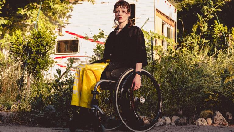 A person sitting in a wheelchair poses in front of an RV trailer parked behind shrubbery.