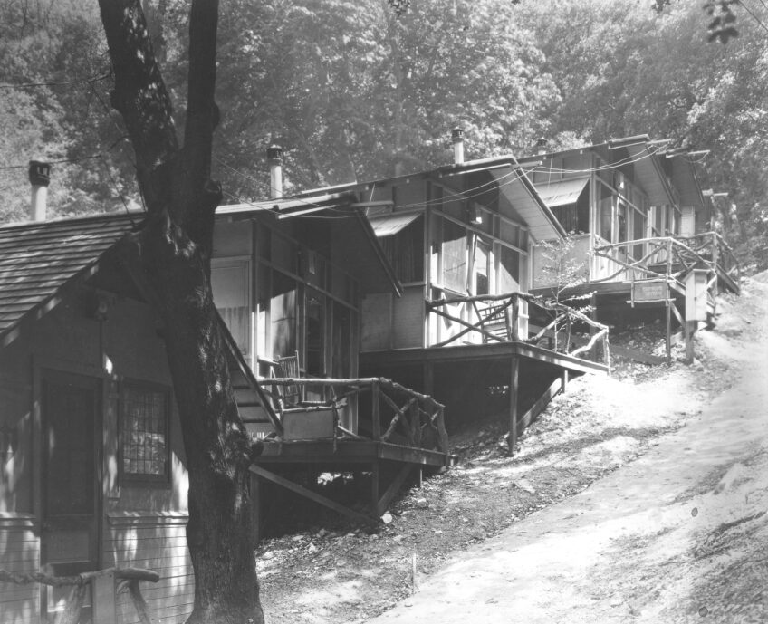 A black and white photo depicts a row of cabins are arranged in a line along a steep slope. Each one is affixed with screened porches. 