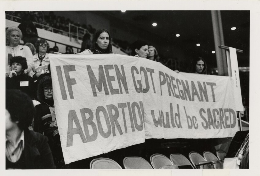 A black and white photo of women holding a banner that reads "If men got pregnant, abortion would be sacred"