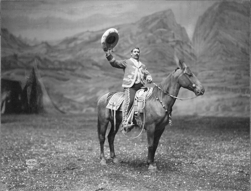 A black and white photo of Vicente Oropeza, a man in distinguished charrería clothing, sitting tall and dignified on a horse as he holds up his hat in the air. He is looking direct to camera. Behind him is a painted Old West landscape.