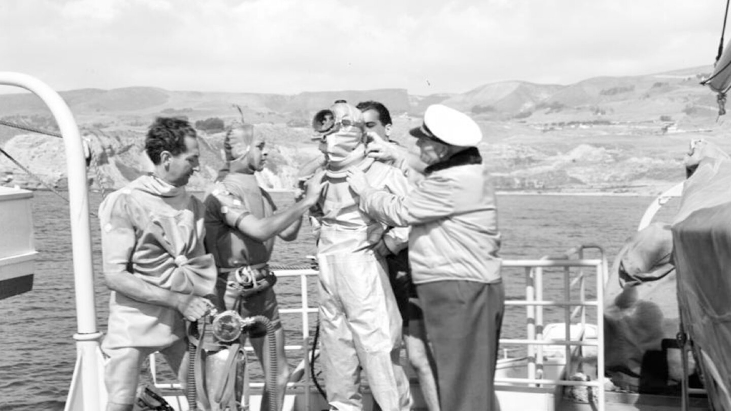Captain Allan Hancock and three other men assist Jacques Cousteau to suit up for scuba diving (aqua-lung diving) to fish for sharks from the Velero IV, ca. 1950. | Allan Hancock Foundation Collection, USC Libraries