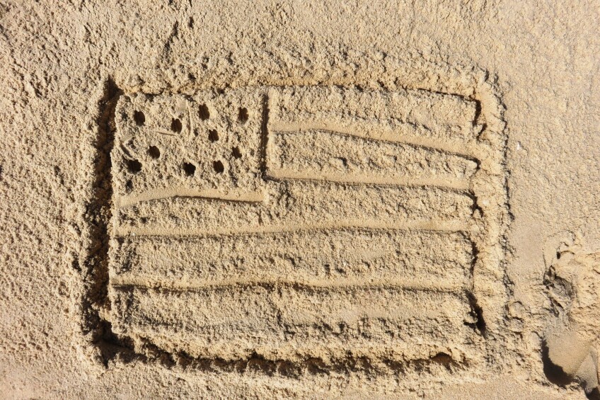 Looking down at sand on a beach, where a simplified drawing of an American flag has been drawn with a stick, featuring 10 round holes representing the 50 stars and five lines representing the 13 stripes.