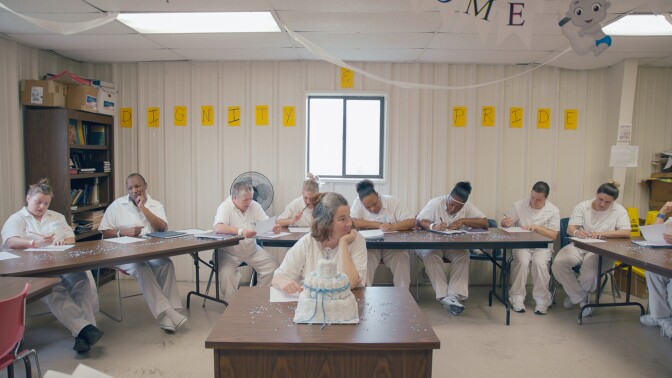 Pregnant inmate Misty sits in the center of a room with a cake celebrating her soon newborn baby in front of her, with fellow inmates sitting around her writing on pieces of paper. | From "America ReFramed: Tutwiler"