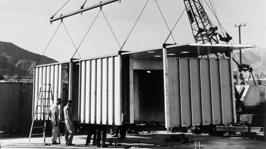 A black and white photo of a steel house under construction. The steel frame of the house is being brought down via a crane.