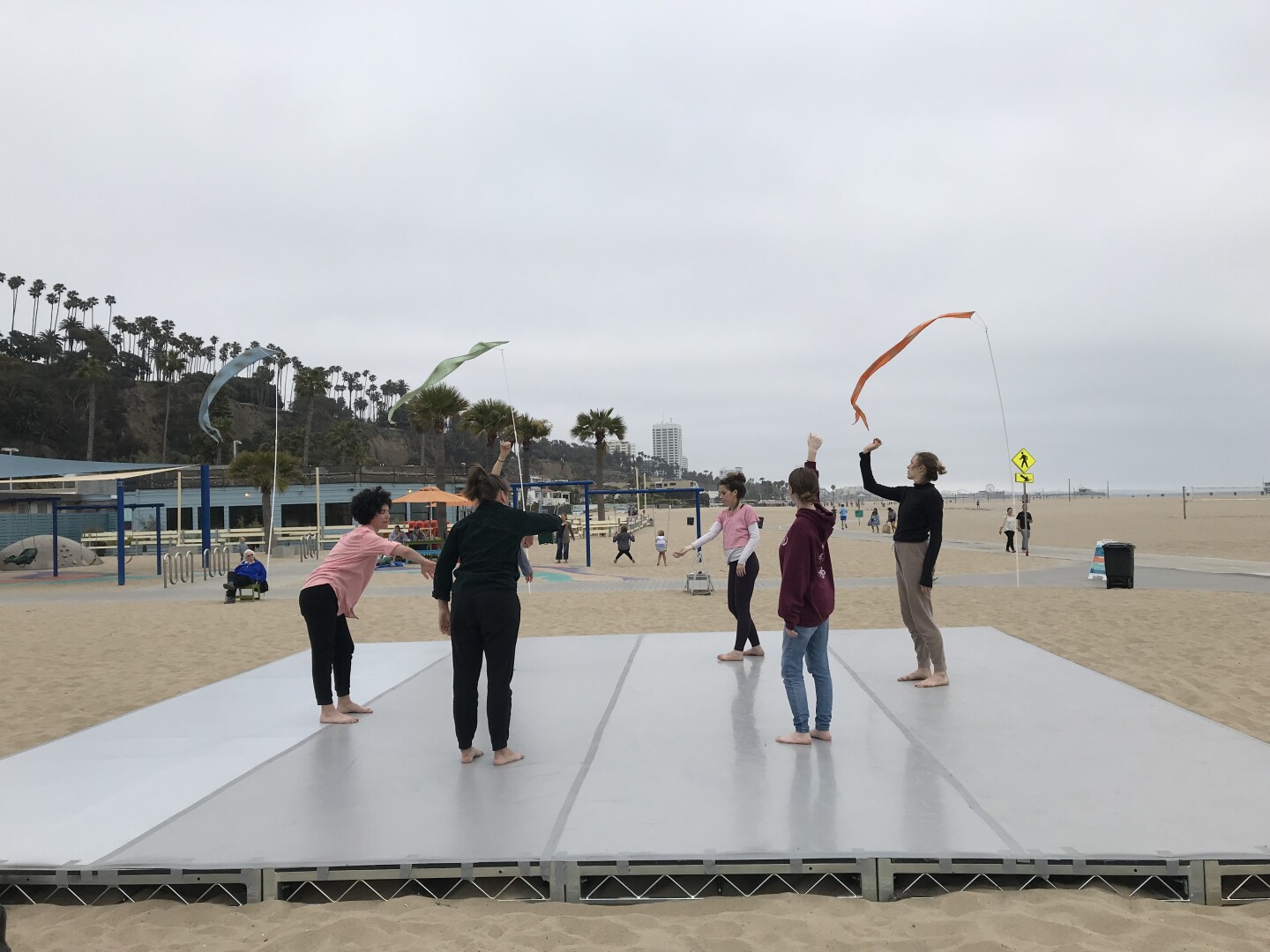 Participants stand on a platform placed on top of the sand at Santa Monica Beach. The participants are waving around different colored scarves in the air. The sky above them is overcast.